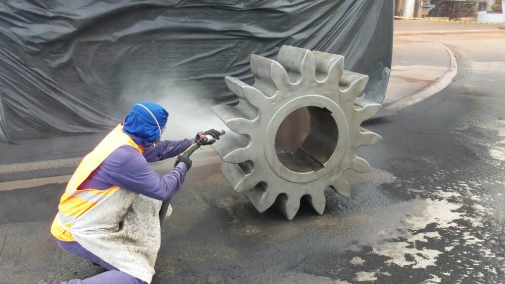 Jatista agachado aplicando jateamento abrasivo úmido em peça industrial no formato de engrenagem com mais ou menos 1 metro de diâmetro